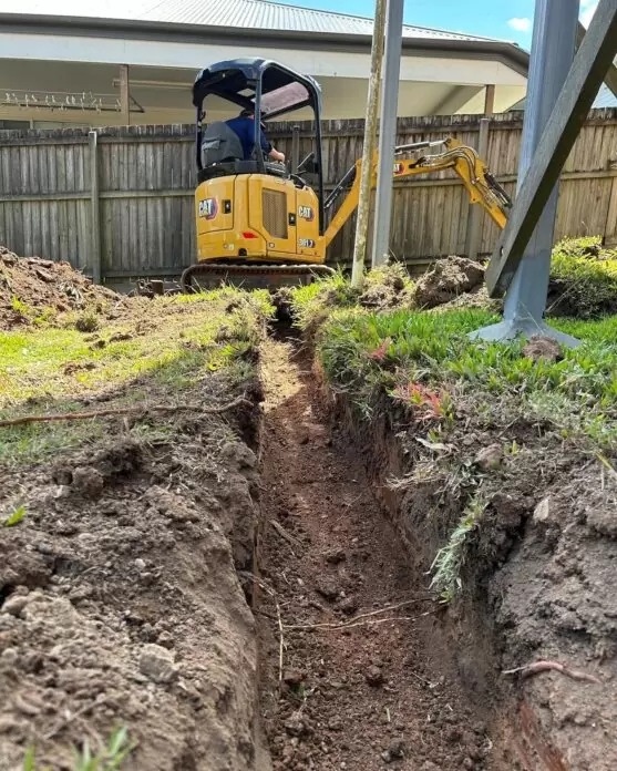 Excavator digging stormwater trench alongside a residential boundary fence in Brisbane