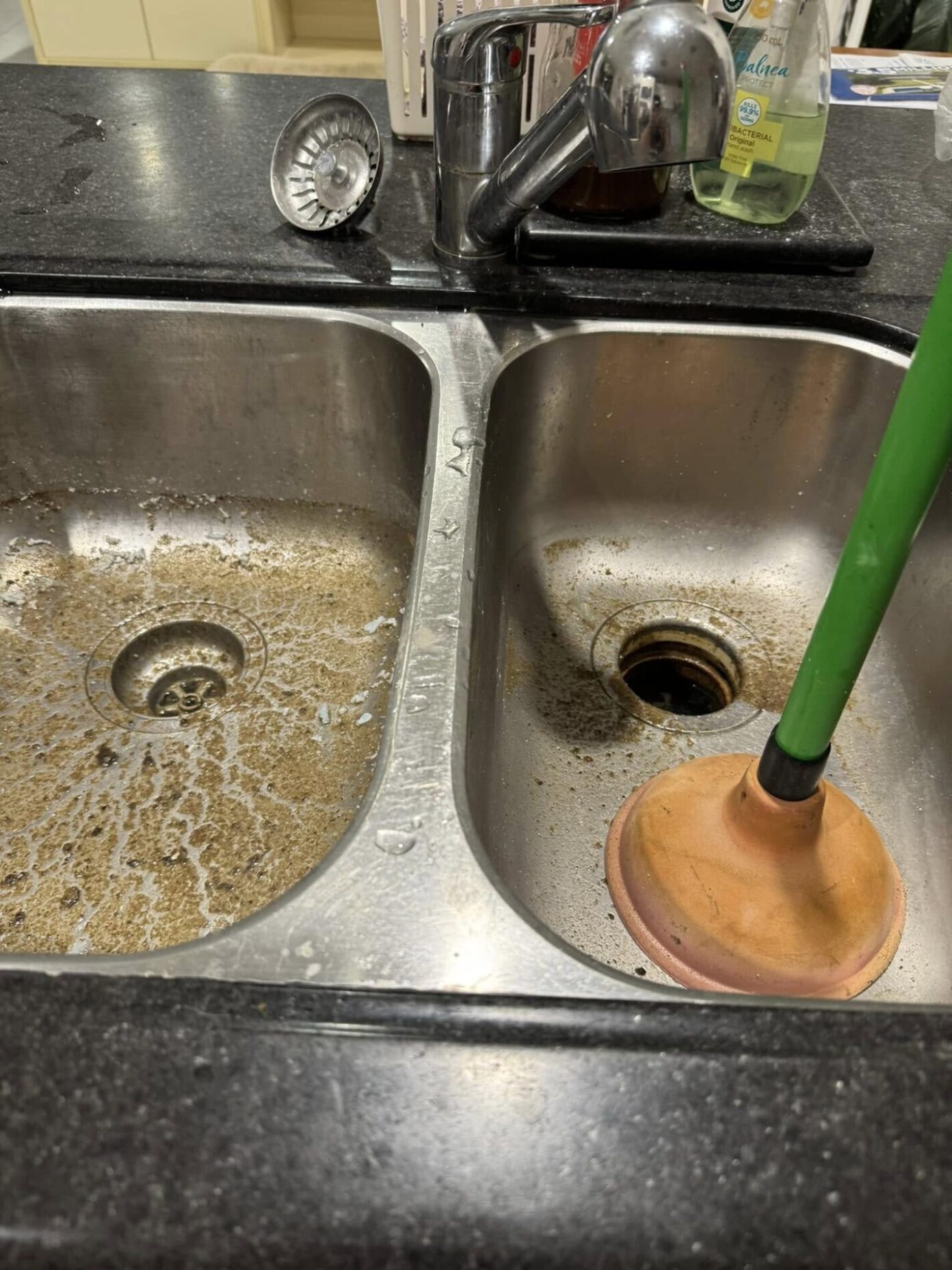 Blocked double kitchen sink with murky water backflow and plunger in use during emergency