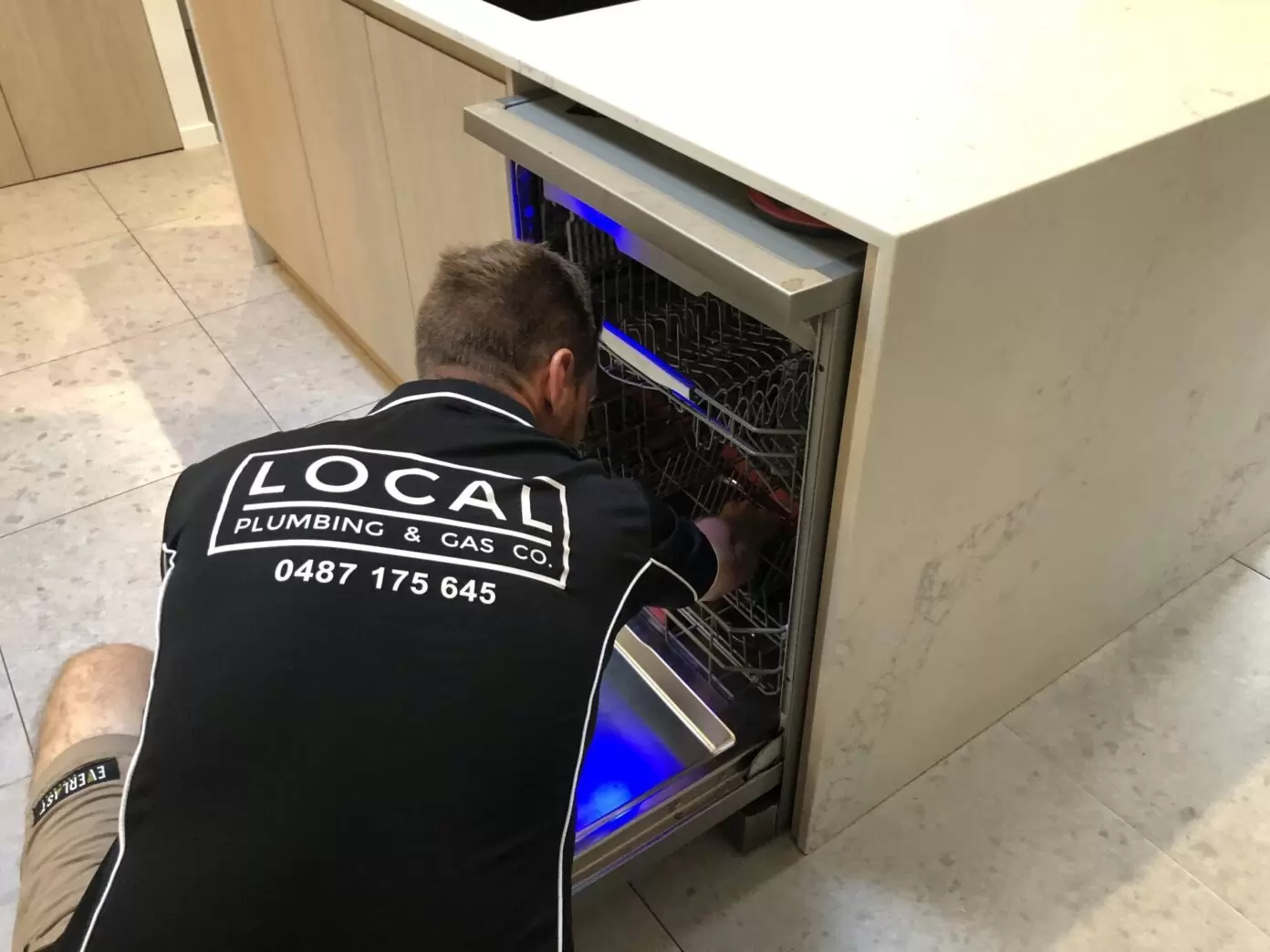 Plumber installing and connecting dishwasher under kitchen bench