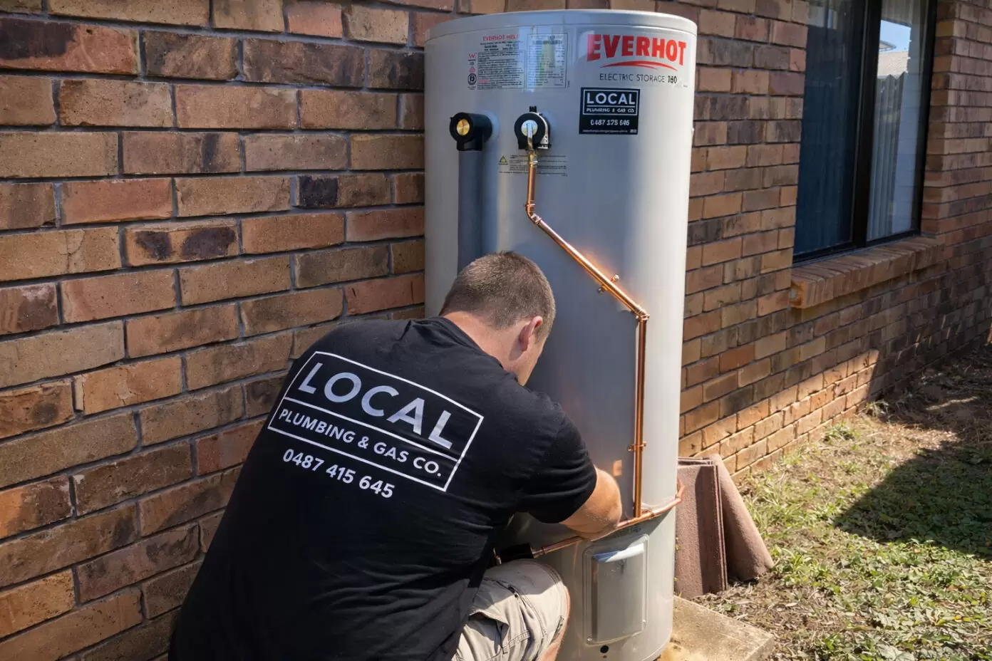 Plumber installing an Everhot electric storage hot water system against a brick wall